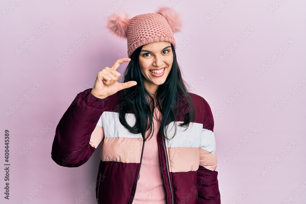 Young hispanic woman wearing wool winter sweater and cap smiling and confident gesturing with hand doing small size sign with fingers looking and the camera. measure concept.