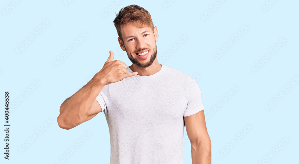 Young caucasian man wearing casual white tshirt smiling doing phone gesture with hand and fingers like talking on the telephone. communicating concepts. Young caucasian man wearing casual white tshirt smiling doing phone gesture with hand and fingers like talking on the telephone. communicating concepts.