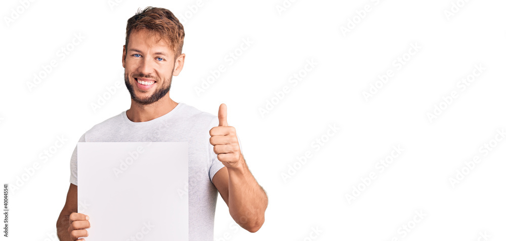 Young caucasian man holding blank empty banner smiling happy and positive, thumb up doing excellent and approval sign Young caucasian man holding blank empty banner smiling happy and positive, thumb up doing excellent and approval sign