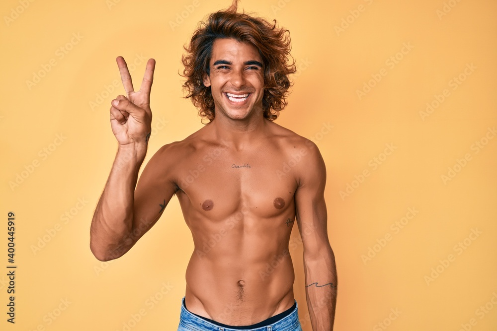 Young hispanic man standing shirtless showing and pointing up with fingers number two while smiling confident and happy.