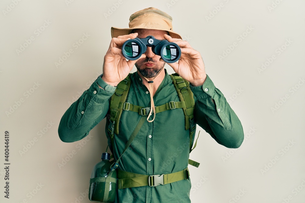 Handsome man with beard wearing explorer hat looking through binoculars puffing cheeks with funny face. mouth inflated with air, catching air. Handsome man with beard wearing explorer hat looking through binoculars puffing cheeks with funny face. mouth inflated with air, catching air.
