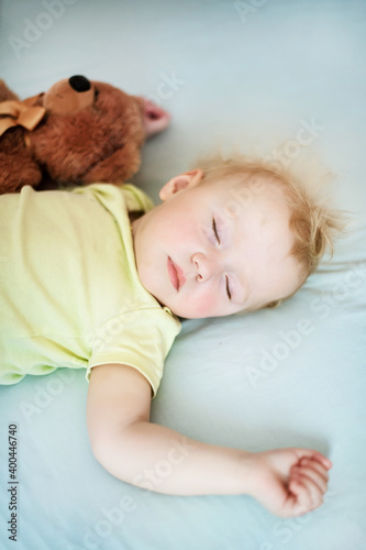 small child sleeps in bed with teddy bear. dishevelled child with blond hair lies closing eyes in bed on blue sheet.