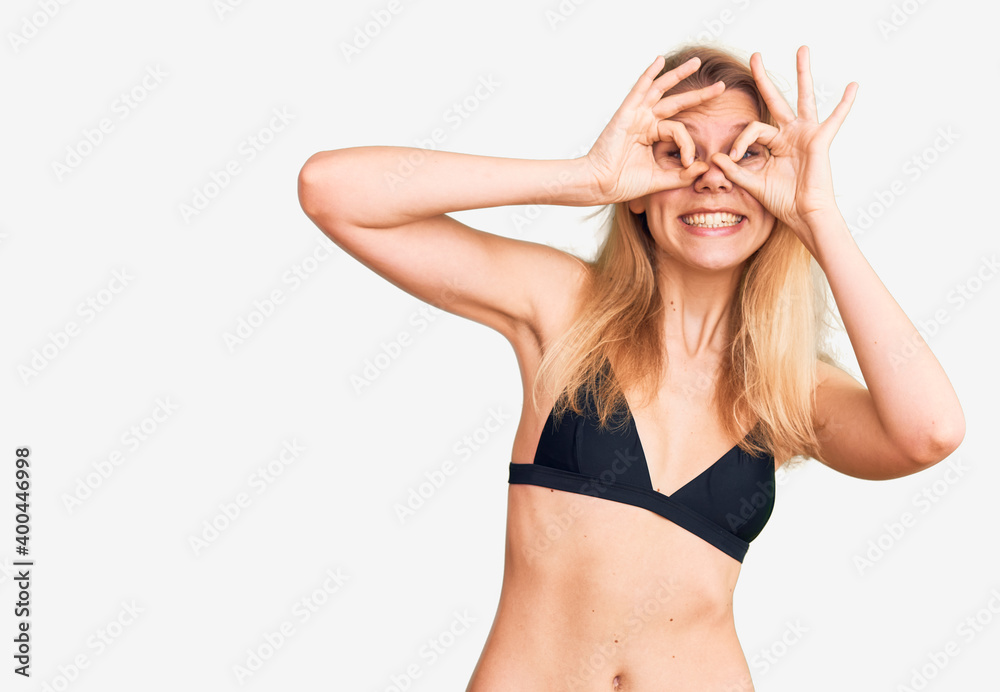 Young beautiful blonde woman wearing bikini doing ok gesture like binoculars sticking tongue out, eyes looking through fingers. crazy expression. Young beautiful blonde woman wearing bikini doing ok gesture like binoculars sticking tongue out, eyes looking through fingers. crazy expression.