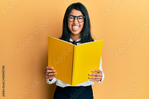 Beautiful asian young woman reading a book wearing glasses sticking tongue out happy with funny expression.