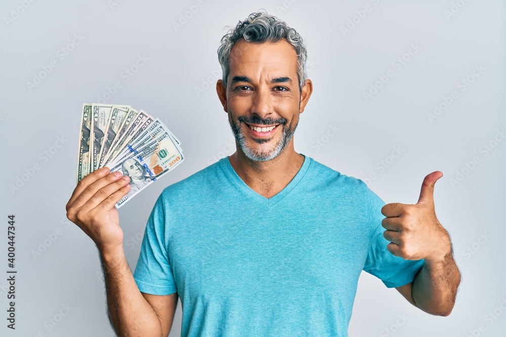Middle age grey-haired man holding dollars smiling happy and positive, thumb up doing excellent and approval sign Middle age grey-haired man holding dollars smiling happy and positive, thumb up doing excellent and approval sign