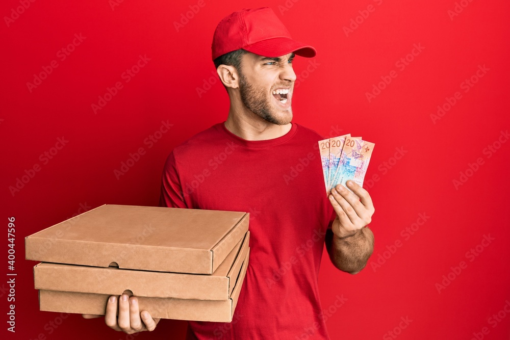 Young caucasian man holding delivery box and swiss franc banknotes angry and mad screaming frustrated and furious, shouting with anger. rage and aggressive concept. Young caucasian man holding delivery box and swiss franc banknotes angry and mad screaming frustrated and furious, shouting with anger. rage and aggressive concept.
