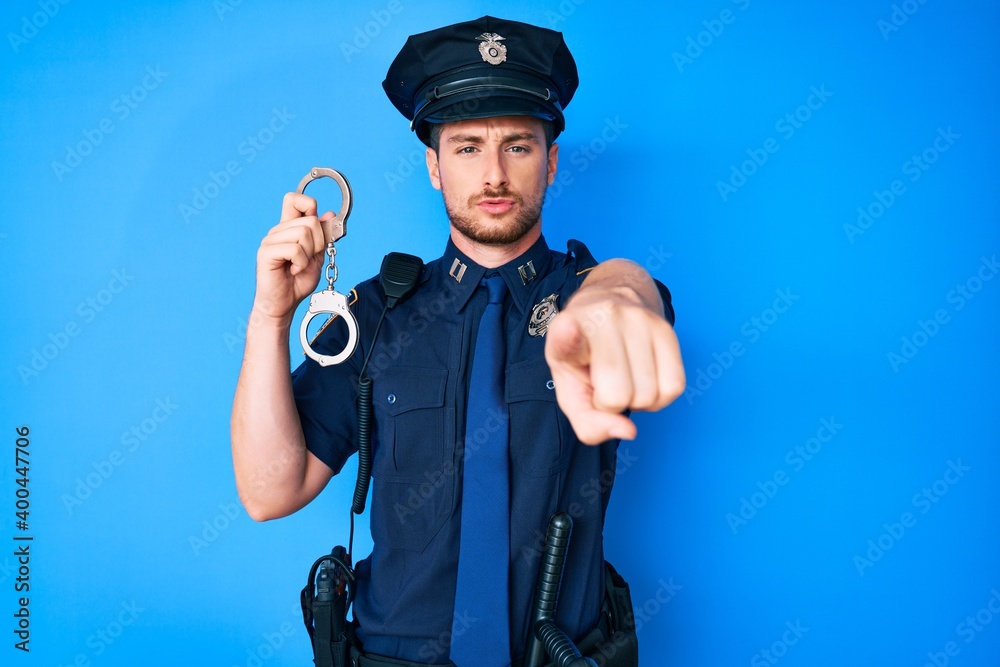 Young caucasian man wearing police uniform holding handcuffs pointing with finger to the camera and to you, confident gesture looking serious Young caucasian man wearing police uniform holding handcuffs pointing with finger to the camera and to you, confident gesture looking serious