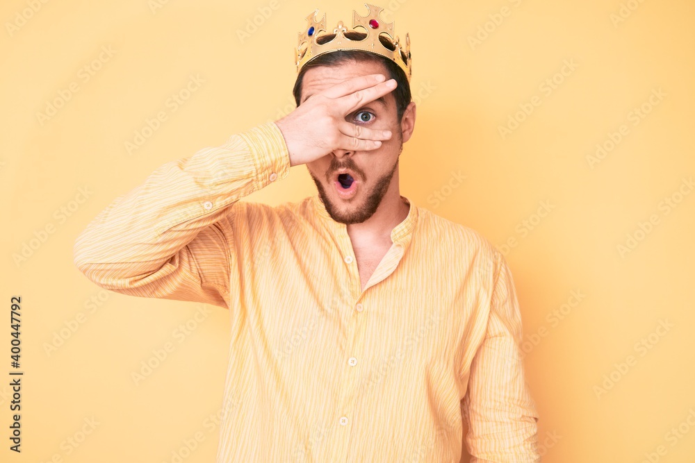 Young handsome man wearing king crown peeking in shock covering face and eyes with hand, looking through fingers afraid Young handsome man wearing king crown peeking in shock covering face and eyes with hand, looking through fingers afraid