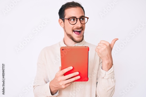 Young handsome man using touchpad device pointing thumb up to the side smiling happy with open mouth