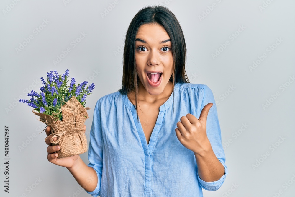 Young hispanic girl holding lavender plant pointing thumb up to the side smiling happy with open mouth Young hispanic girl holding lavender plant pointing thumb up to the side smiling happy with open mouth