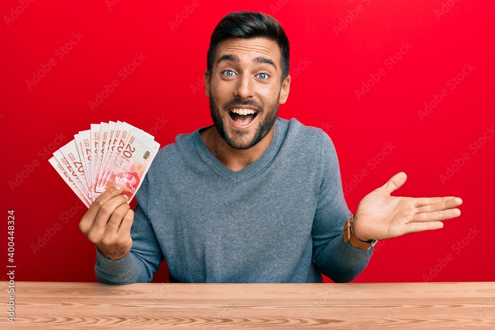 Handsome hispanic man holding israel shekels celebrating achievement with happy smile and winner expression with raised hand Handsome hispanic man holding israel shekels celebrating achievement with happy smile and winner expression with raised hand