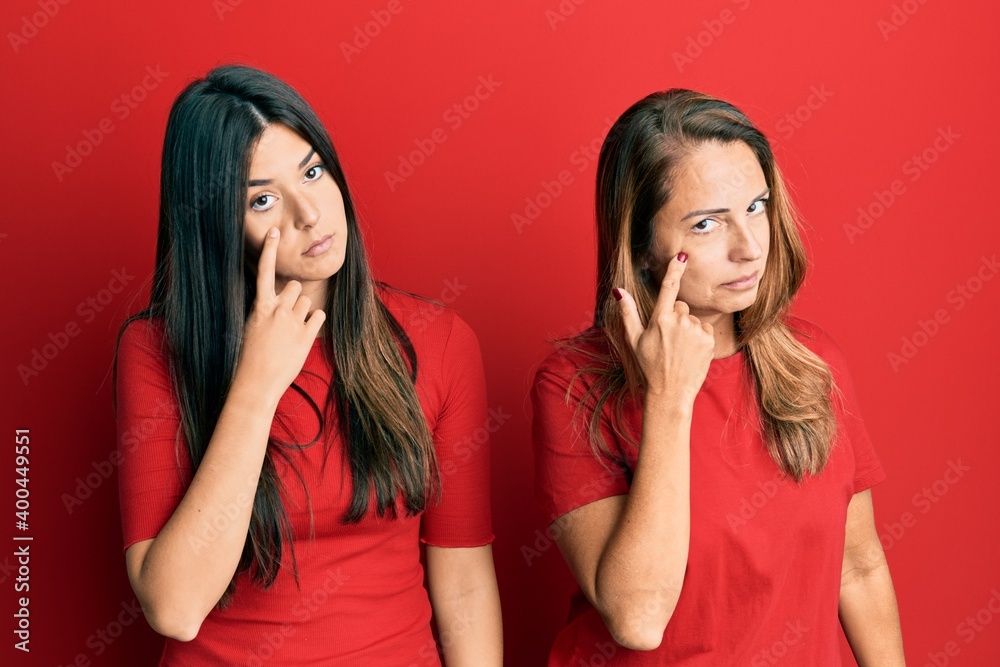 Hispanic family of mother and daughter wearing casual clothes over red background pointing to the eye watching you gesture, suspicious expression Hispanic family of mother and daughter wearing casual clothes over red background pointing to the eye watching you gesture, suspicious expression