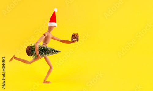 A wooden mannequin in a Christmas hat with a tree under his arm and a gift in his hand.