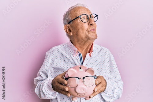 Bild auf Leinwand Senior caucasian man holding piggy bank with glasses smiling looking to the side and staring away thinking