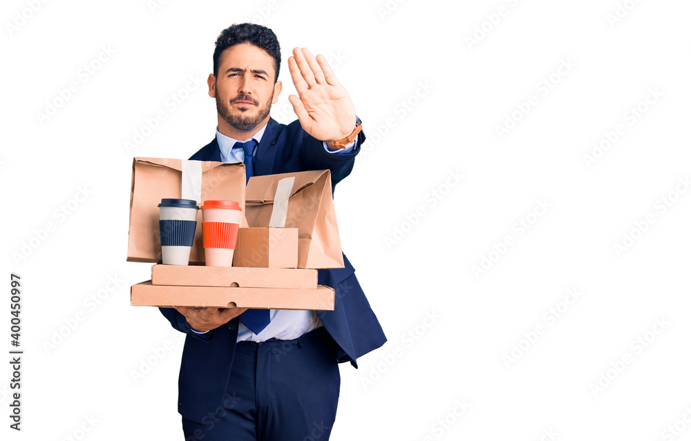 Young hispanic man wearing suit holding takeaway coffee and food with open hand doing stop sign with serious and confident expression, defense gesture Young hispanic man wearing suit holding takeaway coffee and food with open hand doing stop sign with serious and confident expression, defense gesture