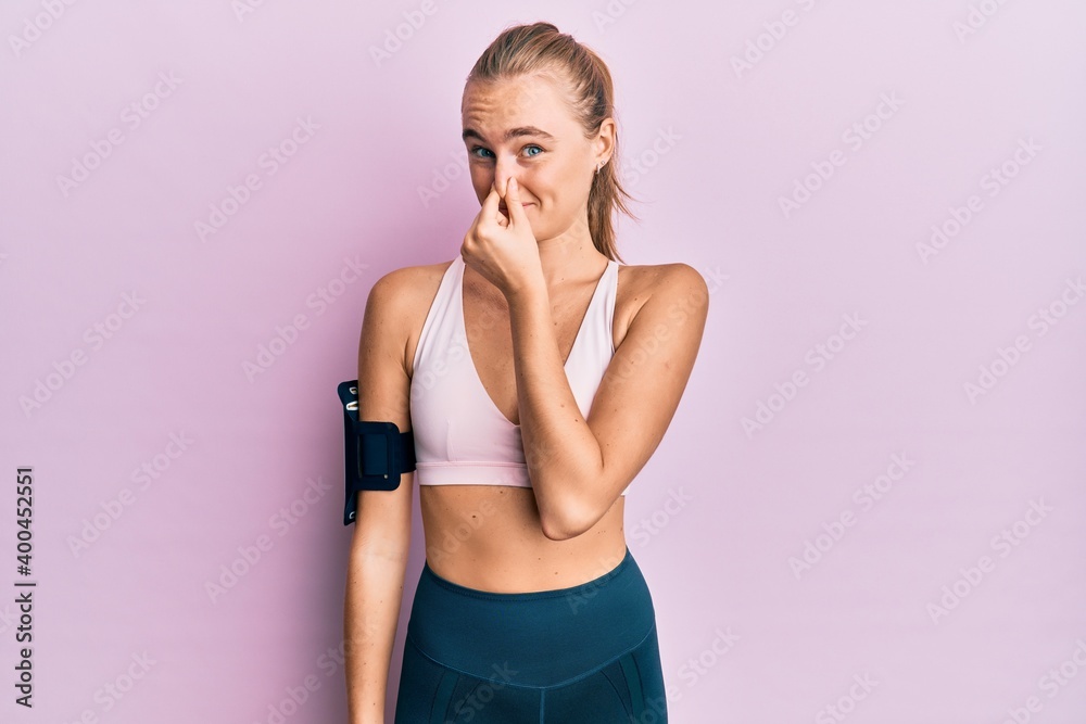 Beautiful blonde woman wearing sportswear and arm band smelling something stinky and disgusting, intolerable smell, holding breath with fingers on nose. bad smell