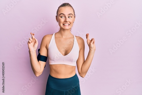 Beautiful blonde woman wearing sportswear and arm band gesturing finger crossed smiling with hope and eyes closed. luck and superstitious concept.