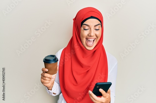 Young beautiful hispanic girl wearing islamic hijab drinking a coffee usign smartphone celebrating crazy and amazed for success with open eyes screaming excited. © Krakenimages.com