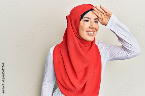 Young beautiful hispanic girl wearing traditional islamic hijab scarf very happy and smiling looking far away with hand over head. searching concept.