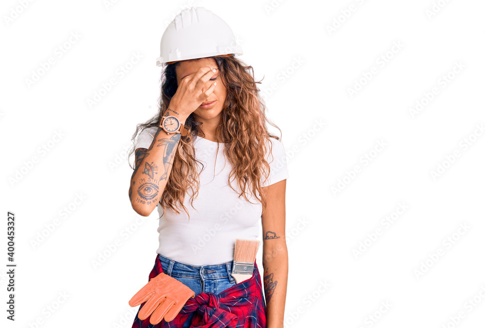 Young hispanic woman with tattoo wearing hardhat and builder clothes tired rubbing nose and eyes feeling fatigue and headache. stress and frustration concept. Young hispanic woman with tattoo wearing hardhat and builder clothes tired rubbing nose and eyes feeling fatigue and headache. stress and frustration concept.
