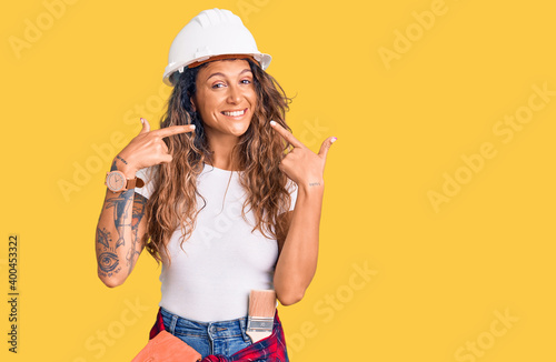 Young hispanic woman with tattoo wearing hardhat and builder clothes smiling cheerful showing and pointing with fingers teeth and mouth. dental health concept.
