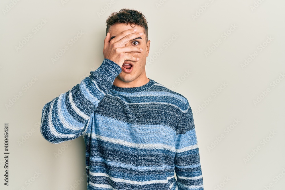 Hispanic young man wearing casual winter sweater peeking in shock covering face and eyes with hand, looking through fingers afraid Hispanic young man wearing casual winter sweater peeking in shock covering face and eyes with hand, looking through fingers afraid