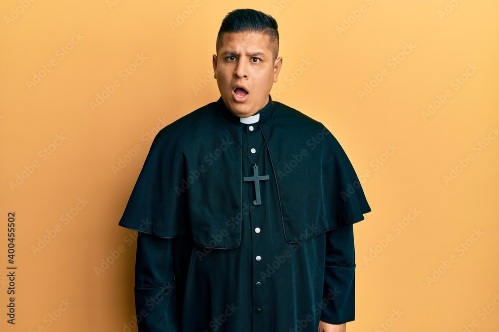 Obraz premium Young latin priest man standing over yellow background in shock face, looking skeptical and sarcastic, surprised with open mouth