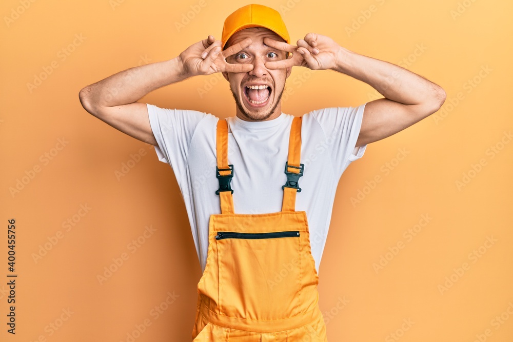 Hispanic young man wearing handyman uniform doing peace symbol with fingers over face, smiling cheerful showing victory Hispanic young man wearing handyman uniform doing peace symbol with fingers over face, smiling cheerful showing victory