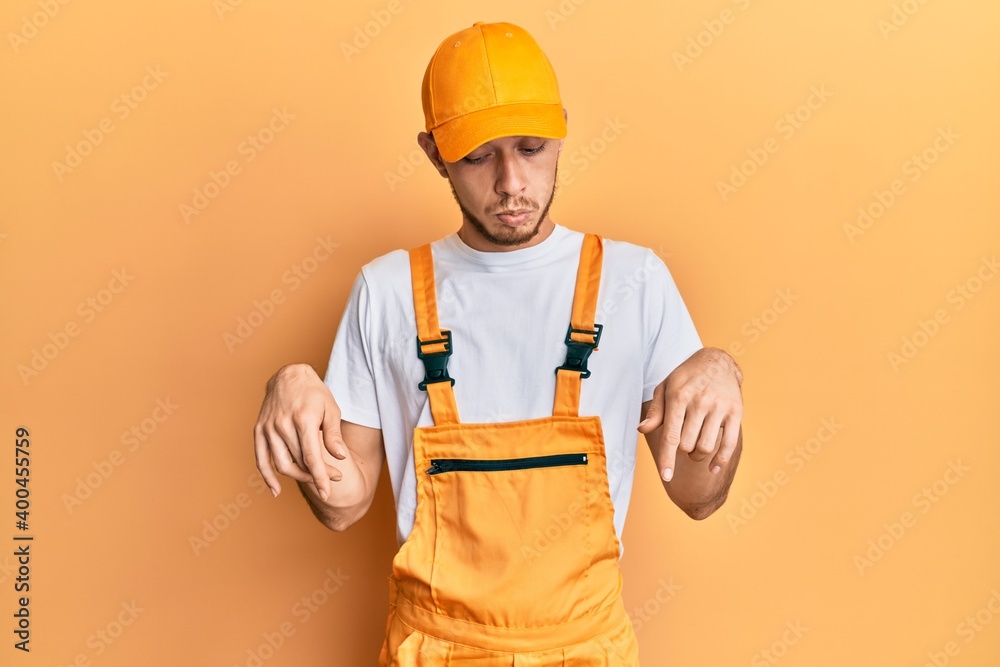 Hispanic young man wearing handyman uniform pointing down looking sad and upset, indicating direction with fingers, unhappy and depressed. Hispanic young man wearing handyman uniform pointing down looking sad and upset, indicating direction with fingers, unhappy and depressed.