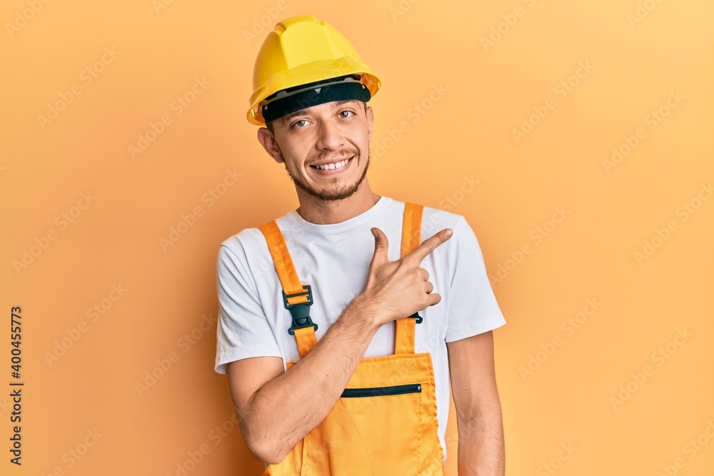 Hispanic young man wearing builder uniform and safety hardhat smiling cheerful pointing with hand and finger up to the side