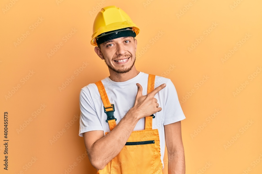Hispanic young man wearing builder uniform and safety hardhat smiling cheerful pointing with hand and finger up to the side Hispanic young man wearing builder uniform and safety hardhat smiling cheerful pointing with hand and finger up to the side