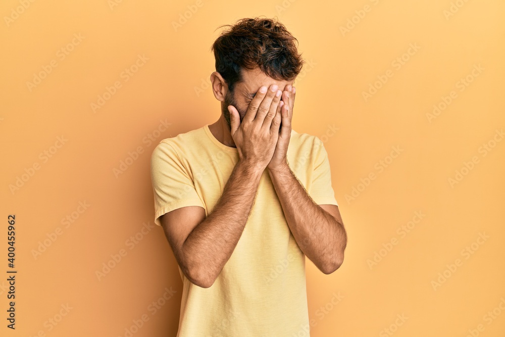 Handsome man with beard wearing casual yellow tshirt over yellow background with sad expression covering face with hands while crying. depression concept. Handsome man with beard wearing casual yellow tshirt over yellow background with sad expression covering face with hands while crying. depression concept.