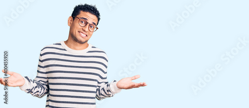 фотографія Hispanic handsome young man wearing casual clothes and glasses clueless and confused expression with arms and hands raised