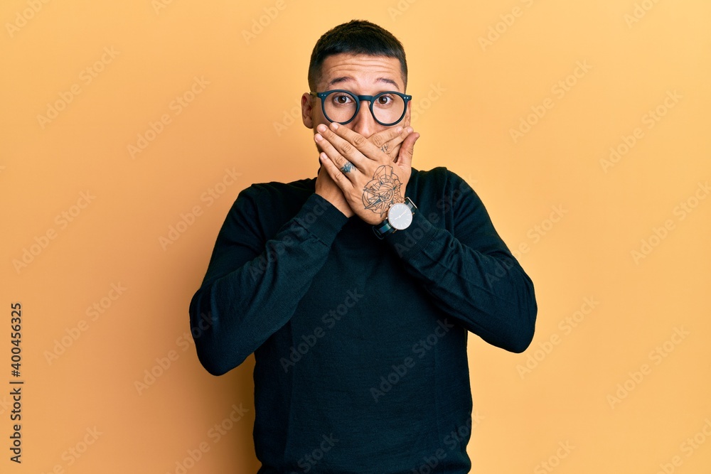 Handsome man with tattoos wearing turtleneck sweater and glasses shocked covering mouth with hands for mistake. secret concept. Handsome man with tattoos wearing turtleneck sweater and glasses shocked covering mouth with hands for mistake. secret concept.