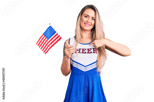 Young beautiful blonde woman wearing cheerleader uniform holding united states flag pointing finger to one self smiling happy and proud
