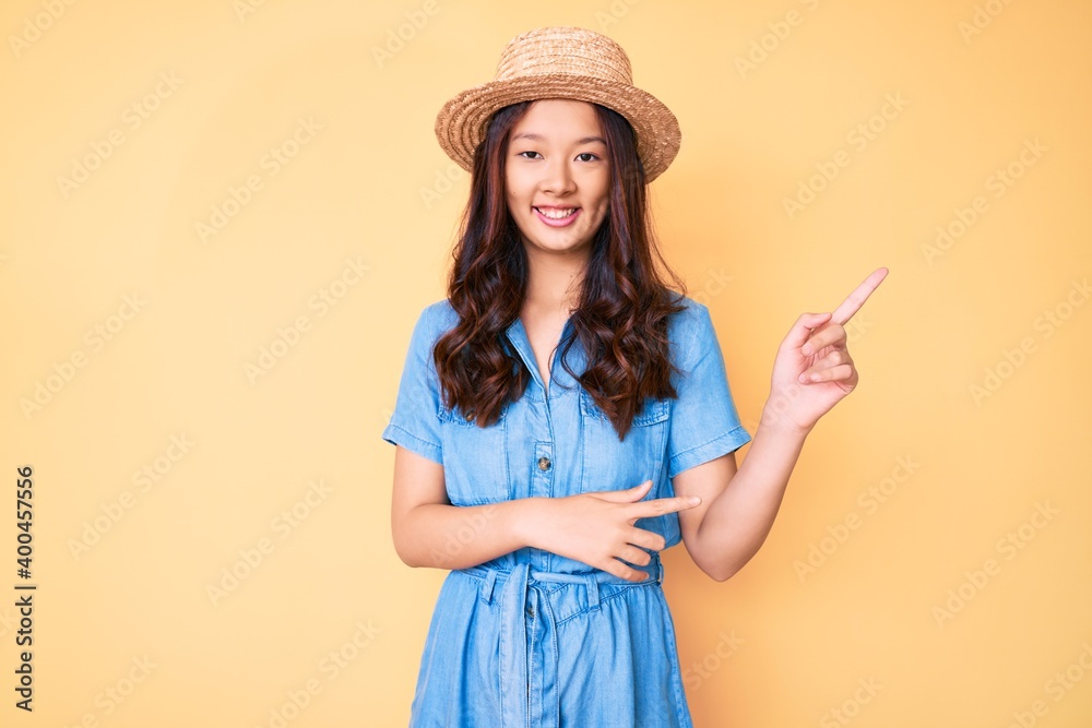 Young beautiful chinese girl wearing summer hat smiling and looking at the camera pointing with two hands and fingers to the side. Young beautiful chinese girl wearing summer hat smiling and looking at the camera pointing with two hands and fingers to the side.