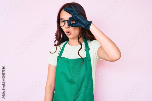 Young beautiful chinese girl wearing cleaner apron doing ok gesture shocked with surprised face, eye looking through fingers. unbelieving expression.
