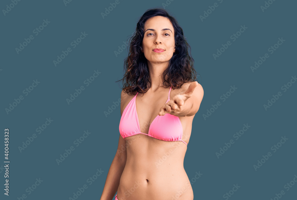 Young beautiful hispanic woman wearing bikini smiling cheerful offering palm hand giving assistance and acceptance. Young beautiful hispanic woman wearing bikini smiling cheerful offering palm hand giving assistance and acceptance.