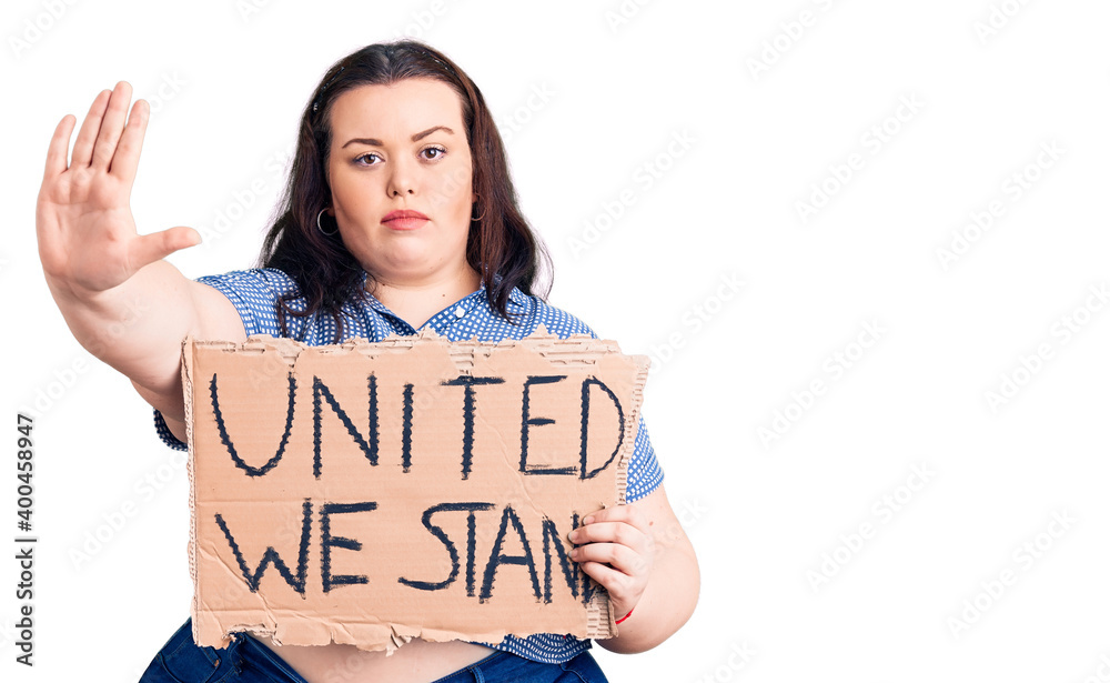 Young plus size woman holding united we stand banner with open hand doing stop sign with serious and confident expression, defense gesture Young plus size woman holding united we stand banner with open hand doing stop sign with serious and confident expression, defense gesture