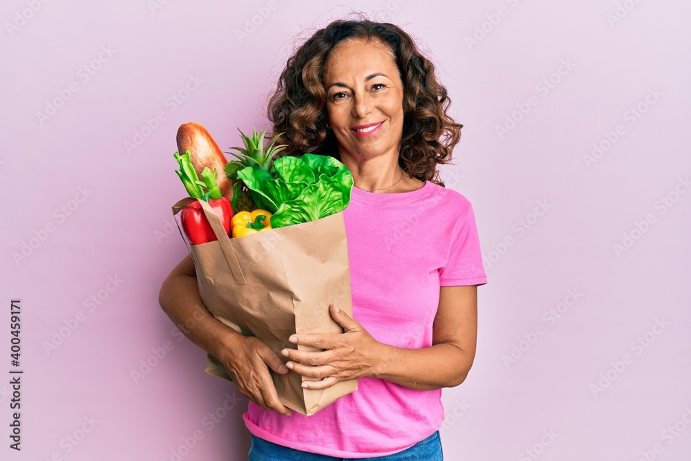 Middle age hispanic woman holding paper bag with bread and groceries looking positive and happy standing and smiling with a confident smile showing teeth Middle age hispanic woman holding paper bag with bread and groceries looking positive and happy standing and smiling with a confident smile showing teeth