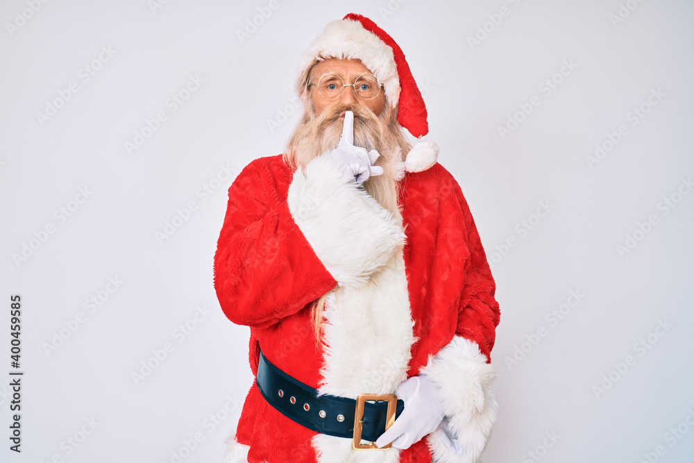 Old senior man with grey hair and long beard wearing santa claus costume with suspenders asking to be quiet with finger on lips. silence and secret concept.