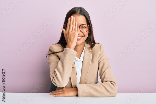 Beautiful hispanic woman working at the office covering one eye with hand, confident smile on face and surprise emotion.
