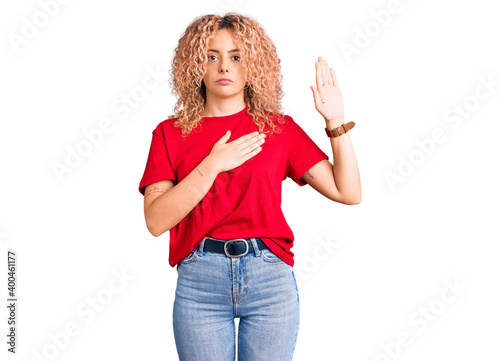 Young blonde woman with curly hair wearing casual red tshirt swearing with hand on chest and open palm, making a loyalty promise oath