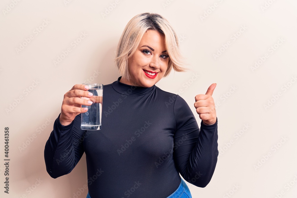 Young beautiful blonde plus size woman drinking glass of water over isolated white background smiling happy and positive, thumb up doing excellent and approval sign Young beautiful blonde plus size woman drinking glass of water over isolated white background smiling happy and positive, thumb up doing excellent and approval sign