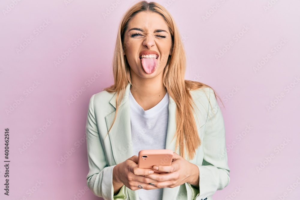 Young caucasian woman wearing business clothes using smartphone sticking tongue out happy with funny expression. Young caucasian woman wearing business clothes using smartphone sticking tongue out happy with funny expression.