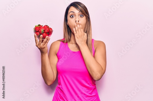 Young beautiful woman holding bowl with strawberries covering mouth with hand, shocked and afraid for mistake. surprised expression