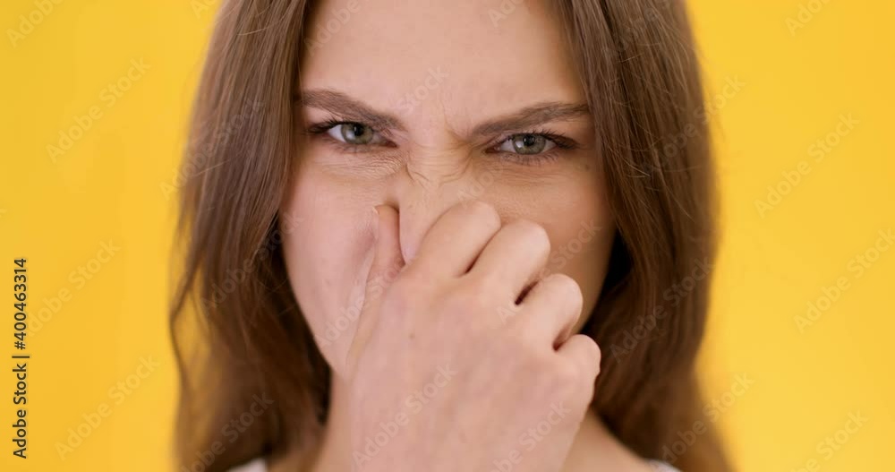 Close up portrait of young woman smelling something awful, closing nose, frowning in displeasure, orange background