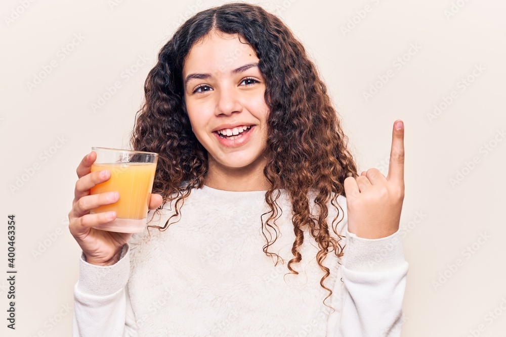 Beautiful kid girl with curly hair drinking glass of orange juice smiling happy pointing with hand and finger to the side