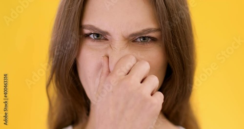 Close up portrait of young woman smelling something awful, closing nose, frowning in displeasure, orange background