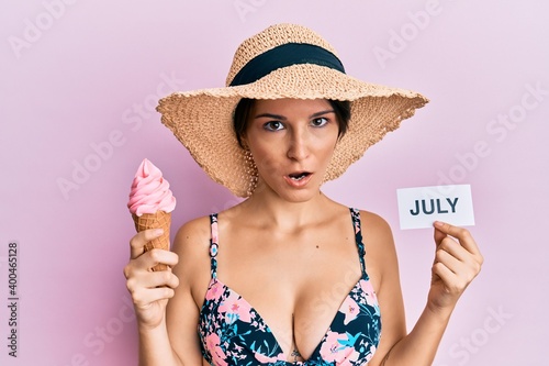 Young brunette woman with short hair holding ice cream and july paper in shock face, looking skeptical and sarcastic, surprised with open mouth